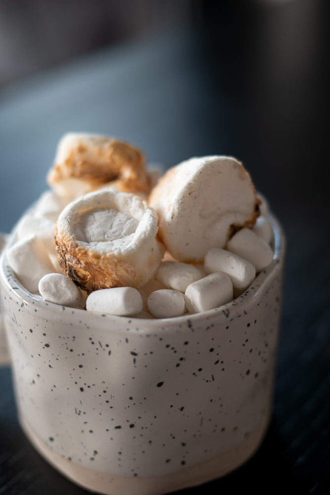 A close-up shot of a speckled mug filled with marshmallows. Some marshmallows are toasted and browned on top, while others are white and fluffy.