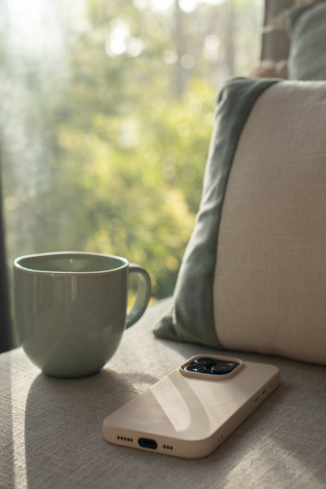 A light green mug sits next to a beige smartphone on a textured gray surface. Sunlight streams in from the left, casting shadows. In the background, a blurred green and white pillow rests against a window with soft bokeh lights.