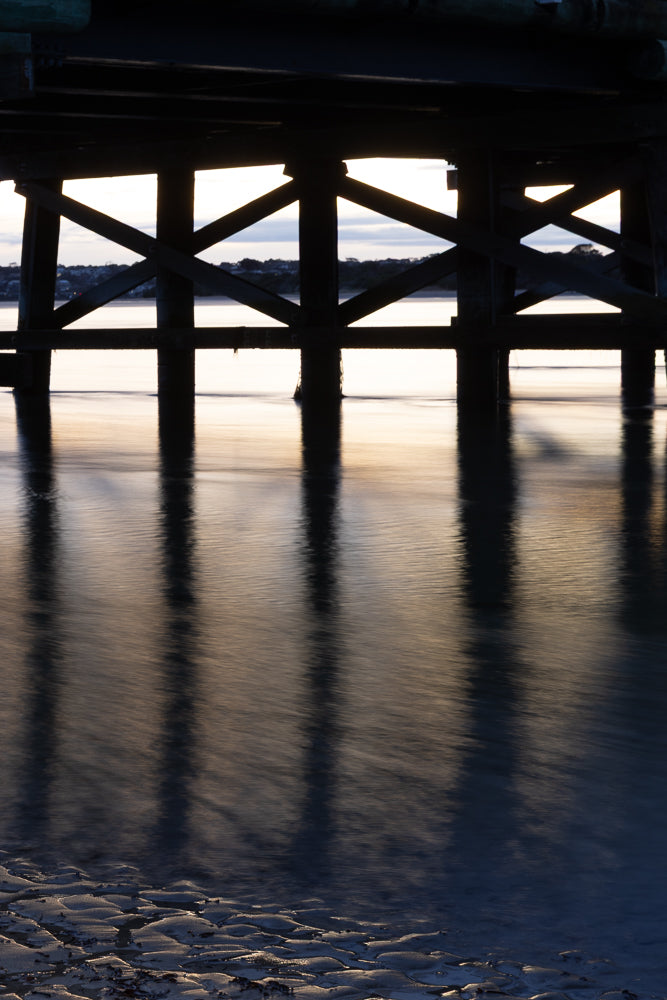 Silhouetted wooden pier supports and their reflections on the water's surface at sunset. The water is calm, creating smooth reflections, and the foreground shows the textured sand of the beach.