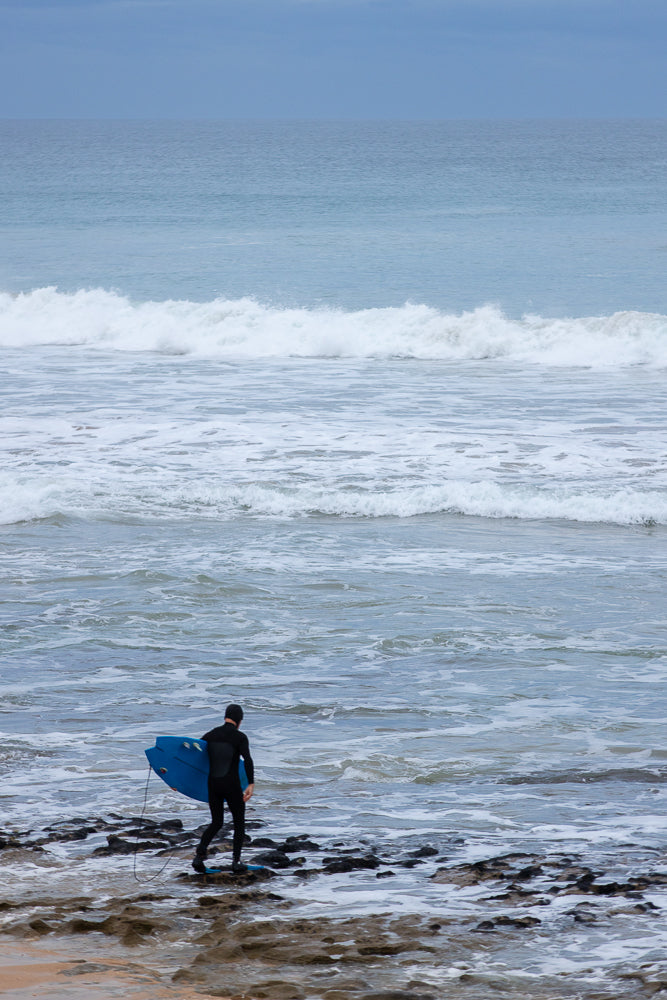 A surfer in a black wetsuit walks into the ocean carrying a blue surfboard. Waves crash on the rocky shore.