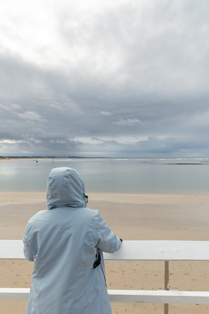 A person wearing a light blue hooded jacket stands on a white railing, looking out at a calm sea under a cloudy sky. The beach in the foreground has footprints.