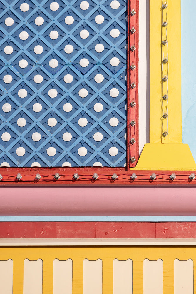 A close-up of a building facade with bold, colorful geometric patterns. The top section features a blue diamond lattice with white circles. Below this is a red band with a string of clear light bulbs. A pink band follows, then a red band, and finally a yellow section with white cutouts.