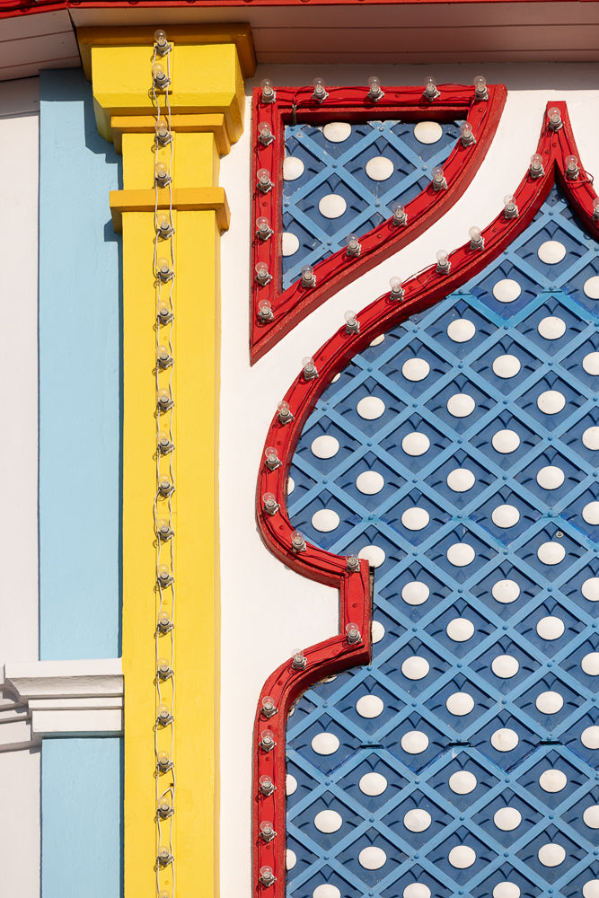 A close-up shot of a building facade with bright yellow, light blue, and white sections. A string of clear light bulbs runs vertically along the edge of a red, ornate frame that borders a blue diamond-patterned section with white circles.