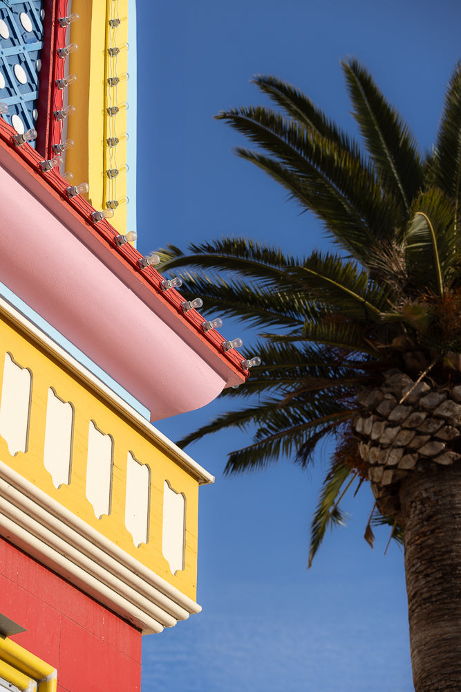 A close-up shot of a colorful building with yellow, pink, and red sections, adorned with a string of lights. A palm tree is visible against a clear blue sky.