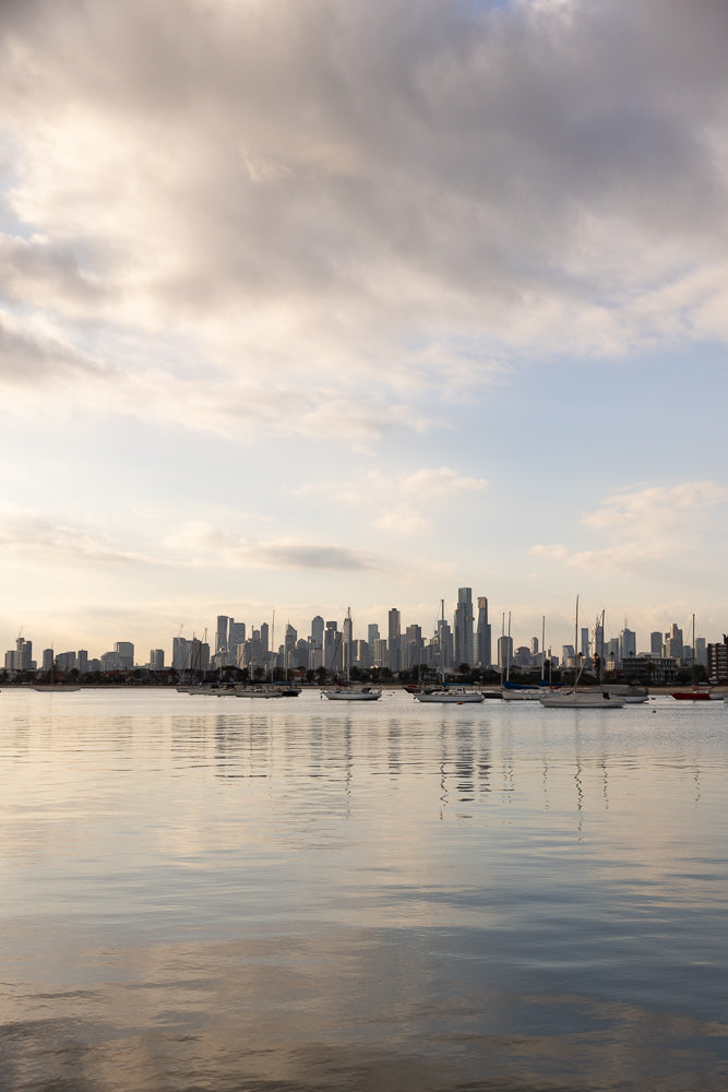 A wide shot of a city skyline across a body of water with sailboats. The sky is partly cloudy with the sun setting or rising, casting a warm glow on the scene.