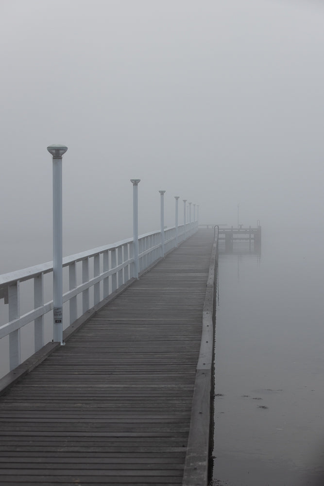 A wooden pier with white railings and lights stretches into a thick fog over calm water.