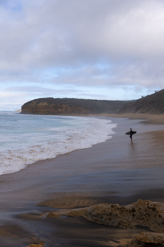 A lone surfer walks along a wet sandy beach carrying a surfboard. Waves crash on the shore, and a large cliff face covered in vegetation rises in the background under a cloudy sky.