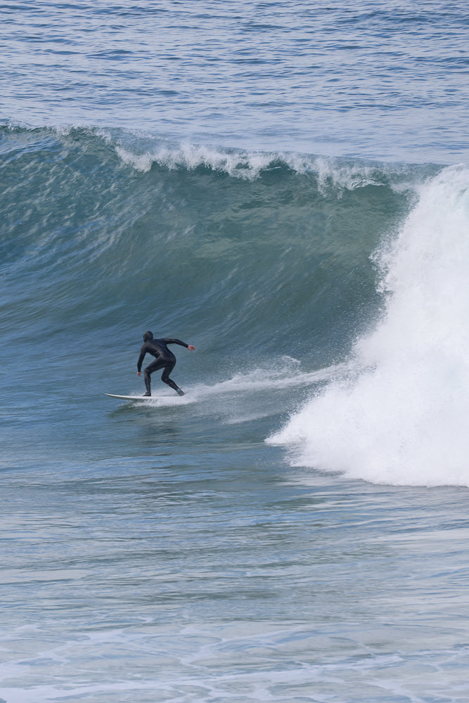 A surfer in a black wetsuit rides a wave on a white surfboard. The surfer is positioned on the left side of the frame, carving through the water as a large wave breaks to their right.