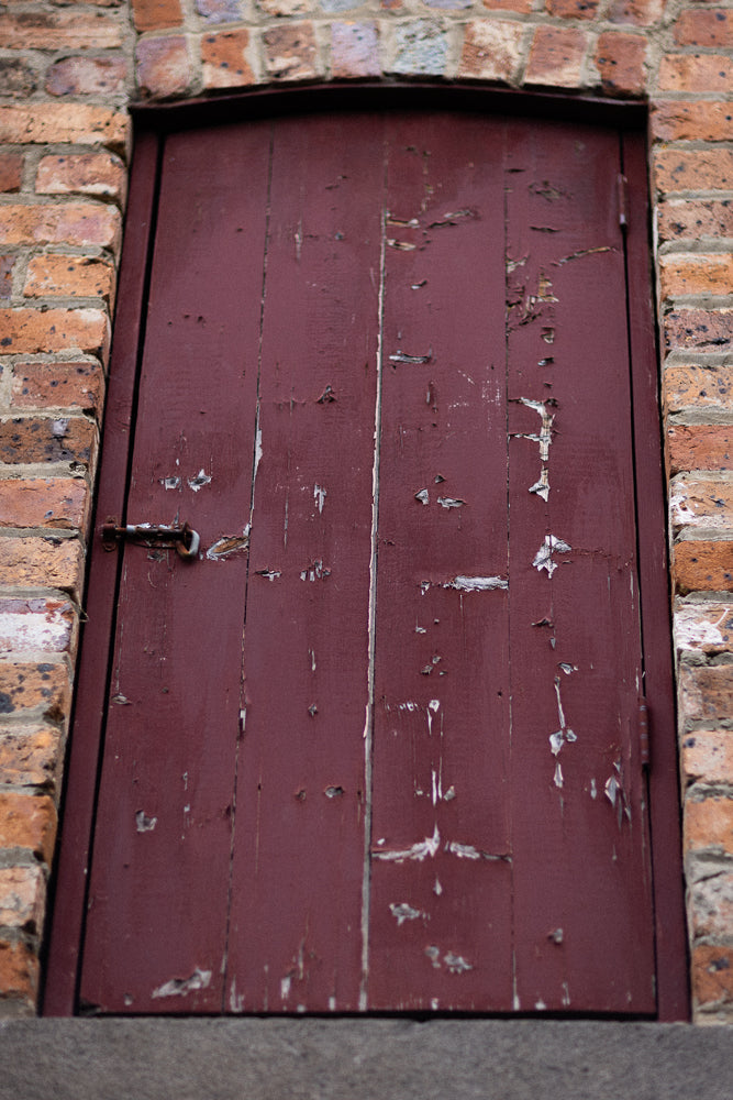 A weathered, dark red wooden door with peeling paint is set within a brick archway. A rusty metal latch is visible on the left side of the door.
