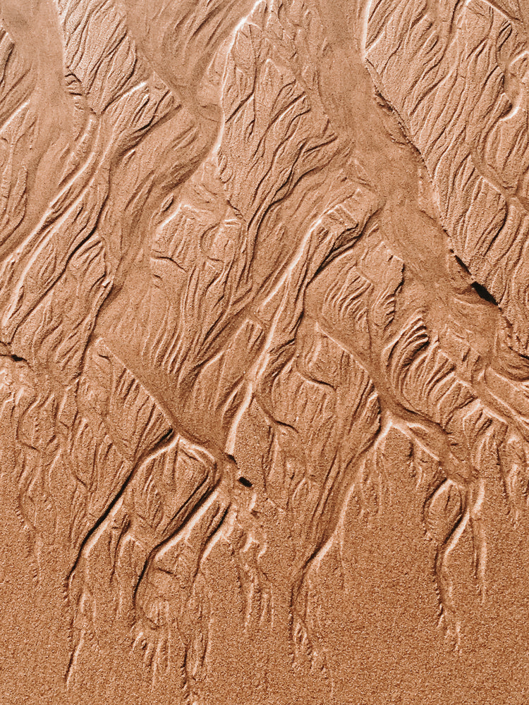 Close-up of wet sand with intricate patterns of ripples and channels formed by receding water. The texture is detailed, showing the granular nature of the sand and the play of light and shadow on the undulating surface.