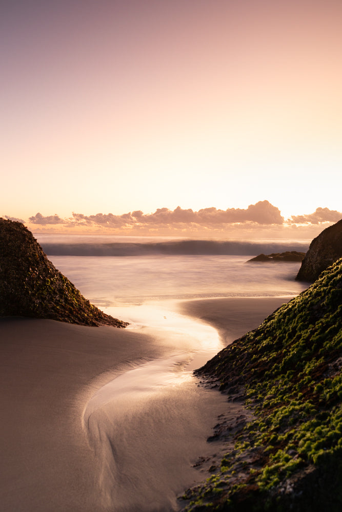 A serene beach scene at sunset, with soft pastel colors in the sky. The gentle waves create a blurred, silky effect on the water. Two moss-covered rocks frame a sandy inlet, reflecting the warm, golden light of the setting sun.