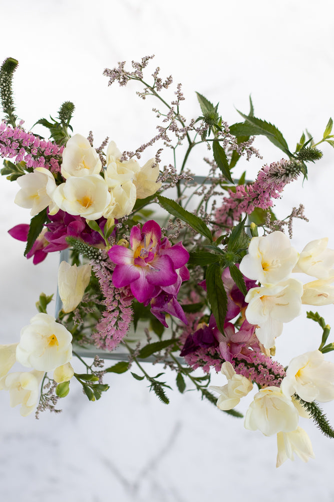 A close-up overhead view of a bouquet of white and pink flowers with green foliage, arranged in a clear glass vase against a white background.