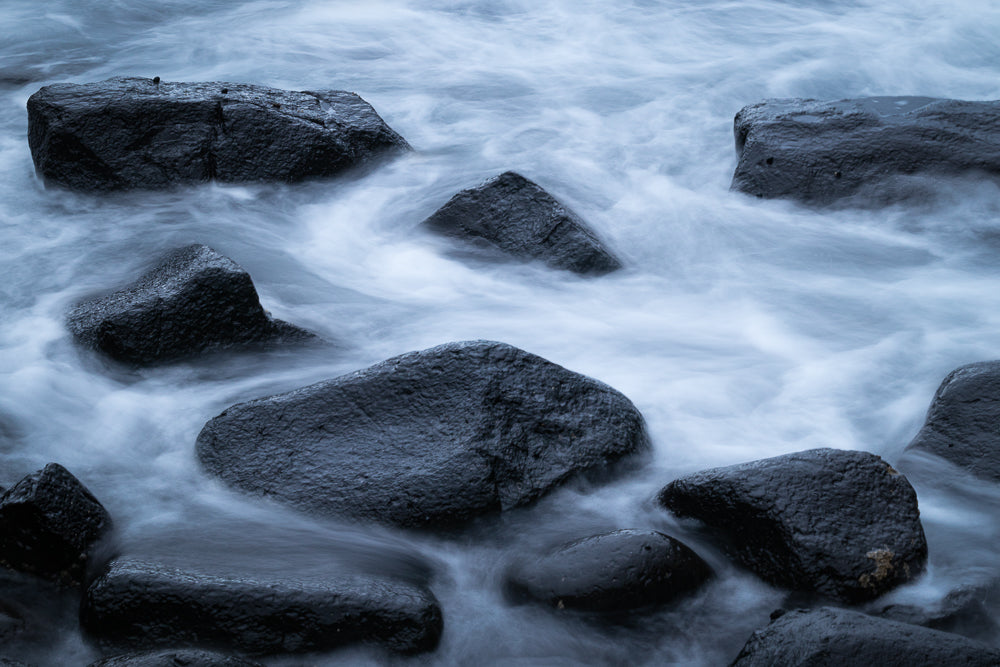 Dark, wet rocks are partially submerged in the ocean, with silky, blurred water flowing around them. The long exposure creates a dreamy, ethereal effect.