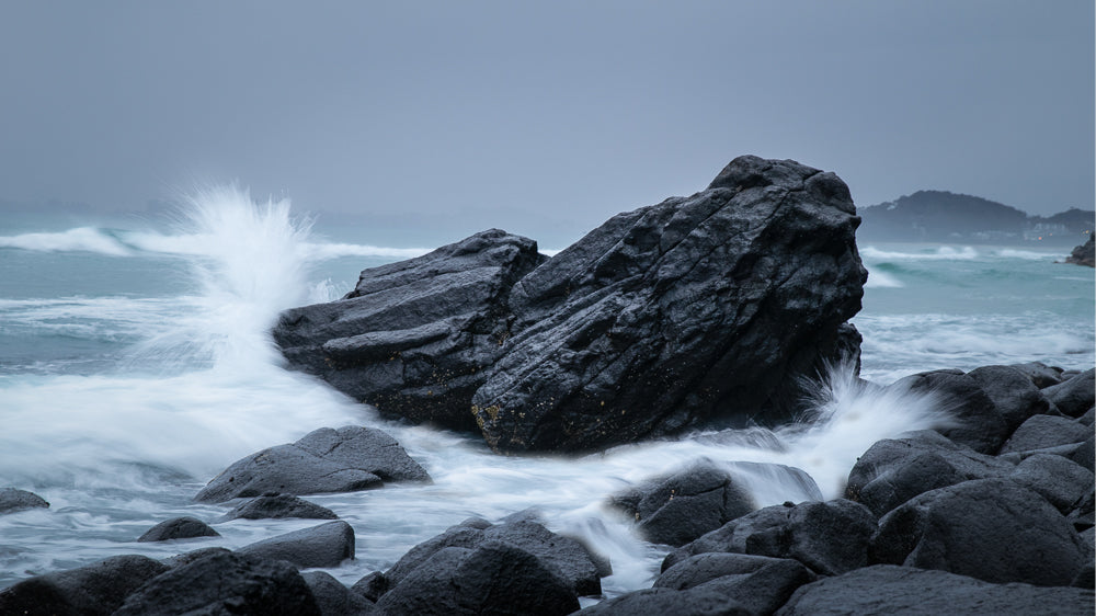 Waves crash against dark, wet rocks on a cloudy day. The water is blurred, creating a sense of motion, while the rocks are sharp and textured. In the distance, a hazy coastline is visible under a muted sky.