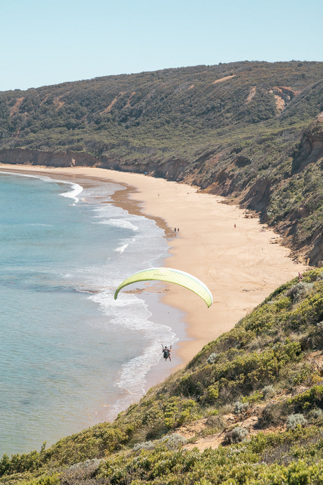 A paraglider with a lime green and white wing soars over a sandy beach with gentle waves. The beach is bordered by a steep, vegetated cliff face under a clear blue sky.