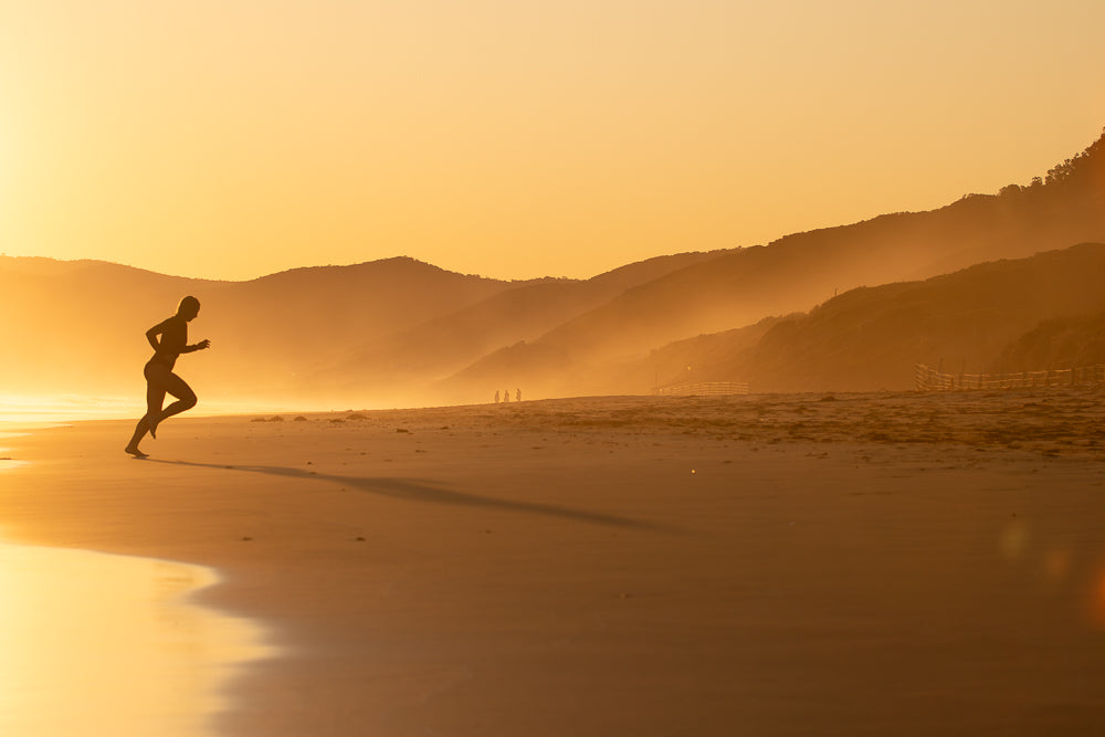 A silhouette of a person running on a sandy beach at sunset. The sky is a warm orange and yellow, with layers of hazy mountains in the background. The runner's shadow stretches long across the sand.