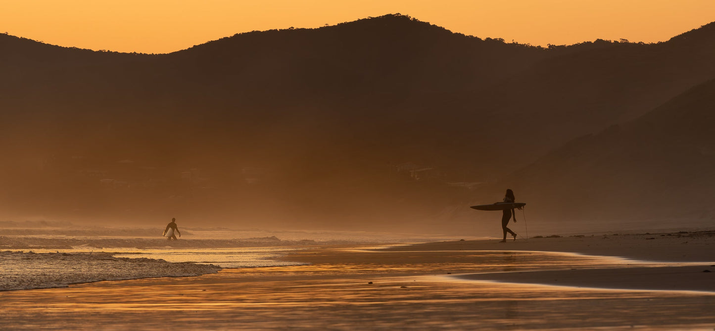 A surfer walks along a beach carrying a surfboard at sunset. The sky is a warm orange, and the mountains in the background are silhouetted against the light. The wet sand reflects the colors of the sunset.