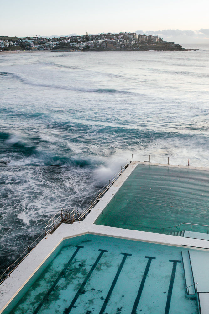 An aerial view of the Bondi Icebergs swimming pool in Sydney, Australia, with waves crashing against the rocks and the ocean stretching to the horizon. The pool is filled with clear blue water and has lane markers visible.