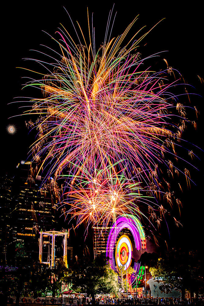 A vibrant fireworks display illuminates the night sky above a city skyline, with a Ferris wheel and carnival rides visible below. The fireworks are a mix of colors, including pink, purple, green, and gold, with streaks of light fanning out across the frame. The city buildings are lit up, and a crowd of people can be seen gathered below, enjoying the spectacle.