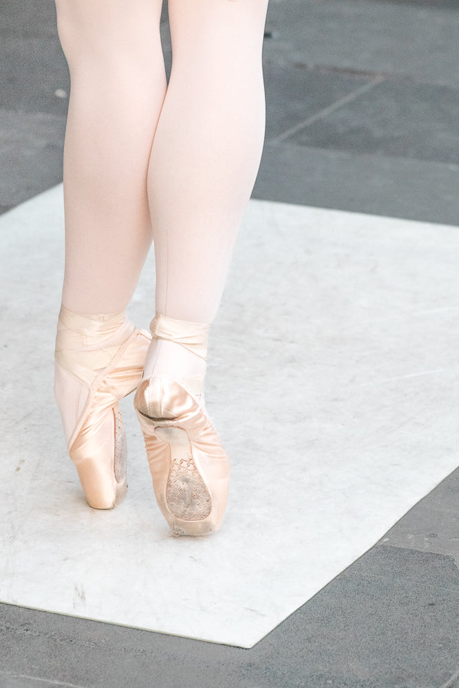 Close-up of a ballet dancer's legs and feet in pink tights and pointe shoes, standing on a white surface with a grey background.