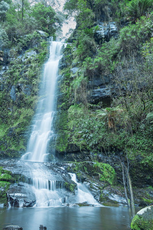 A tall, multi-tiered waterfall cascades down a moss-covered rocky cliff face surrounded by lush green forest and ferns. The water appears silky smooth due to a long exposure, flowing into a calm pool at the bottom.