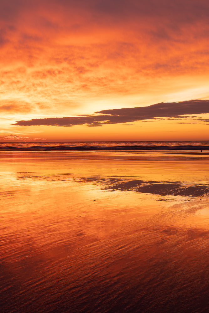 A dramatic sunset paints the sky in fiery oranges and reds, reflected in the wet sand of a beach. The ocean horizon is a thin, dark line separating the vibrant sky from the shimmering shore.