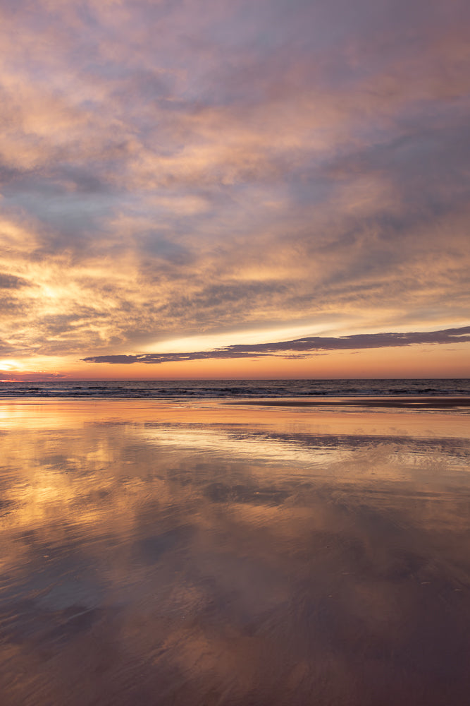 A serene beach scene at sunset. The sky is filled with wispy clouds painted in shades of pink, orange, and purple. The calm ocean reflects the vibrant colors of the sky, creating a mirror-like effect on the wet sand.