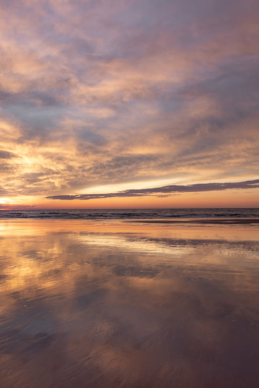 A serene beach scene at sunset. The sky is filled with wispy clouds painted in shades of pink, orange, and purple. The calm ocean reflects the vibrant colors of the sky, creating a mirror-like effect on the wet sand.