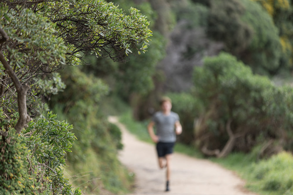 A person runs on a dirt path through a wooded area. Green foliage is in the foreground, and the runner is blurred in the background.