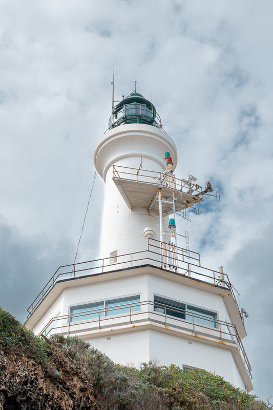 A white lighthouse with a green lantern room stands against a cloudy sky. The lighthouse has multiple levels with railings and lights, including red and green signal lights.
