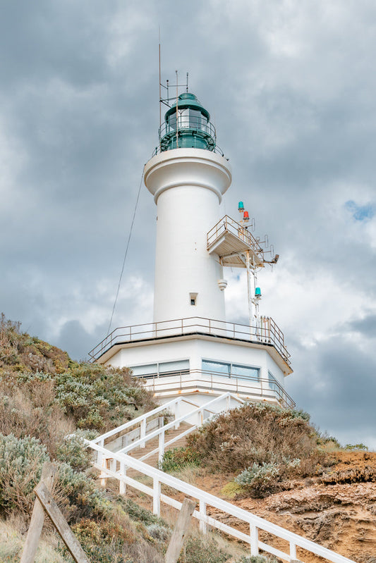 A white lighthouse stands tall on a hill overlooking the ocean, with a cloudy sky above. A white staircase leads up to the lighthouse, surrounded by dry brush and rocks. The lighthouse is equipped with navigational lights and antennas, suggesting its purpose is to guide ships at sea.