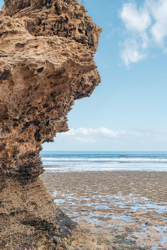 A large, porous rock formation juts out from the left side of the frame, against a backdrop of a calm blue ocean and sky with scattered clouds. The foreground shows a rocky, tide-pooled beach.