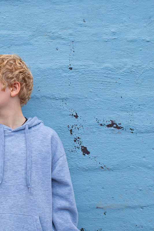 A young boy with curly blonde hair, wearing a light blue hoodie, stands in front of a textured blue wall. The boy is shown from the shoulders up, with his head turned to the left, revealing his ear and the side of his face.