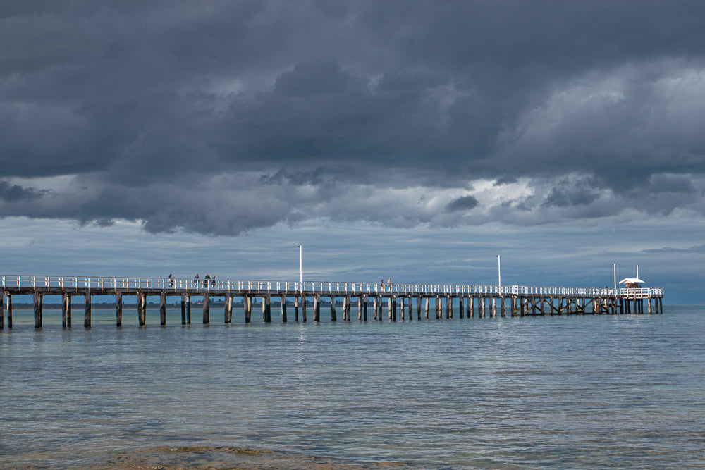 A long wooden pier extends into calm, shallow water under a dramatic, dark, cloudy sky. Several people are visible walking along the pier, which has white railings and wooden pilings.