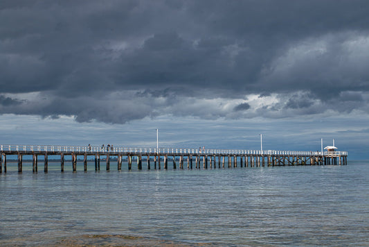 A long wooden pier extends into calm, shallow water under a dramatic, dark, cloudy sky. Several people are visible walking along the pier, which has white railings and wooden pilings.
