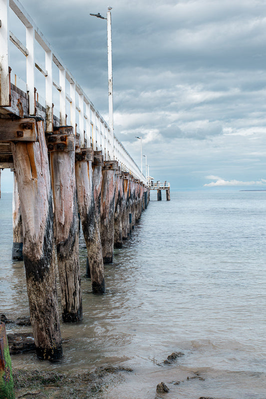 A weathered wooden pier extends into calm water under a cloudy sky. The pier's thick, barnacle-covered pilings are visible, supporting a white railing. A few people are visible at the far end of the pier.