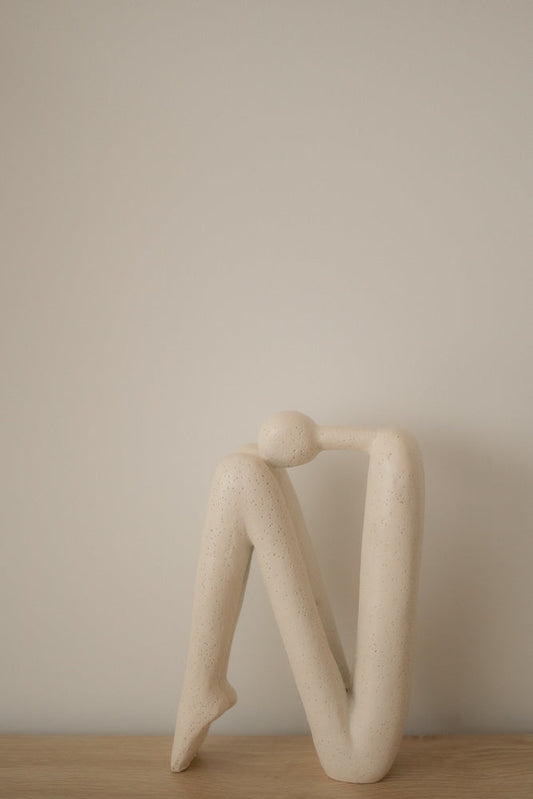 A minimalist, cream-colored abstract sculpture of a seated figure with its head resting on its knees. The sculpture has a textured surface with small dark speckles.