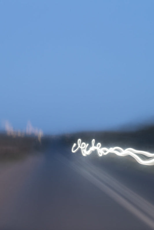 A blurry image of a road at dusk with a bright white light trail curving across the right side of the frame. The sky is a clear, soft blue.