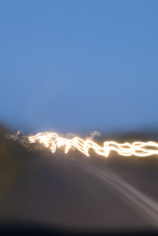 Abstract image of light trails against a blue sky. The bright yellow-white light forms a wavy, serpentine pattern across the lower half of the frame, suggesting movement and speed.