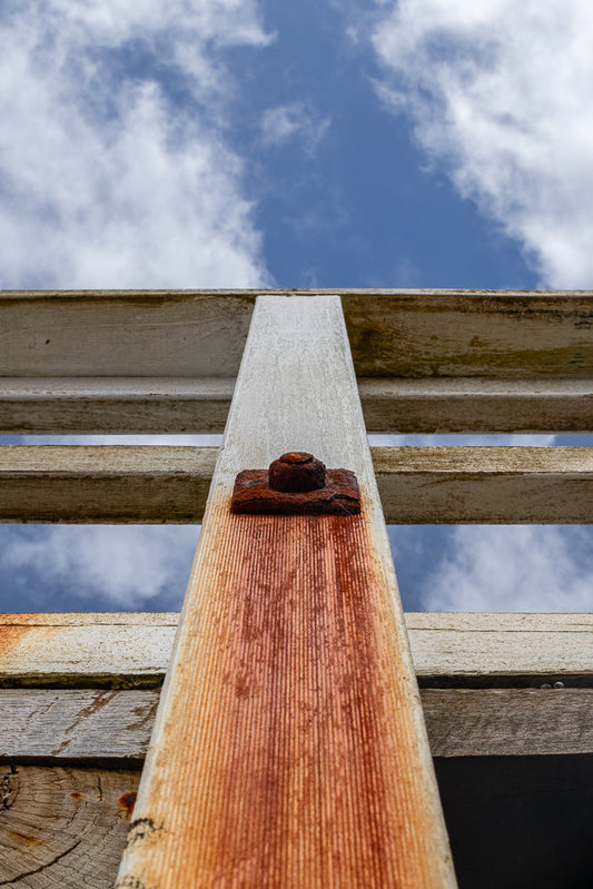 A low-angle shot looking up at a weathered wooden structure against a blue sky with white clouds. A rusty bolt and washer secure a vertical wooden post to horizontal planks.