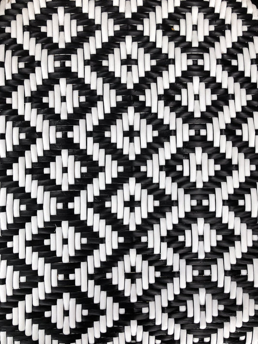 A close-up, overhead view of a woven pattern made of black and white plastic or wicker material. The pattern is a repeating geometric design of diamonds and squares formed by the interlacing strands.