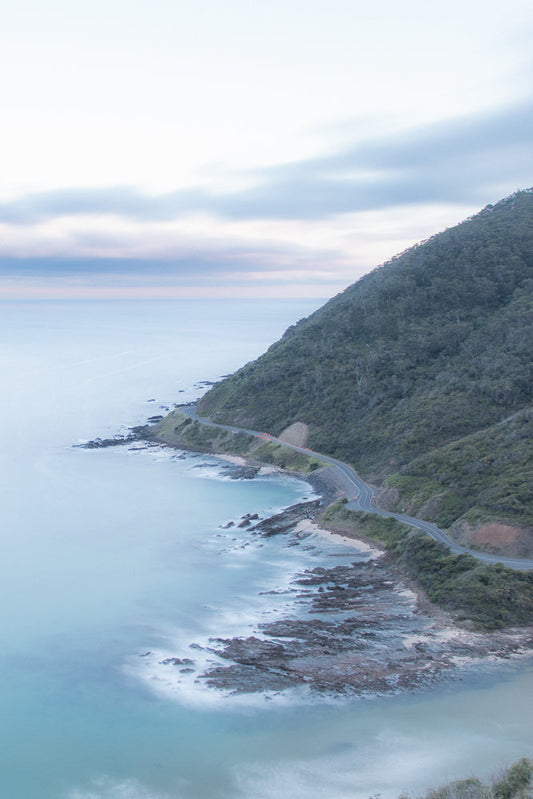 A serene aerial view of Great Ocean Road, clouds and sky at sunset, with soft pink and blue hues blending into the vast expanse.