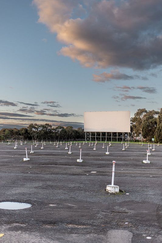 A wide shot of an empty drive-in movie theater parking lot at dusk. The large white screen is visible in the distance, surrounded by trees. Numerous white posts with red tops are spaced out across the asphalt, marking parking spots. The sky above is a mix of blue and dramatic clouds tinged with orange and gray.