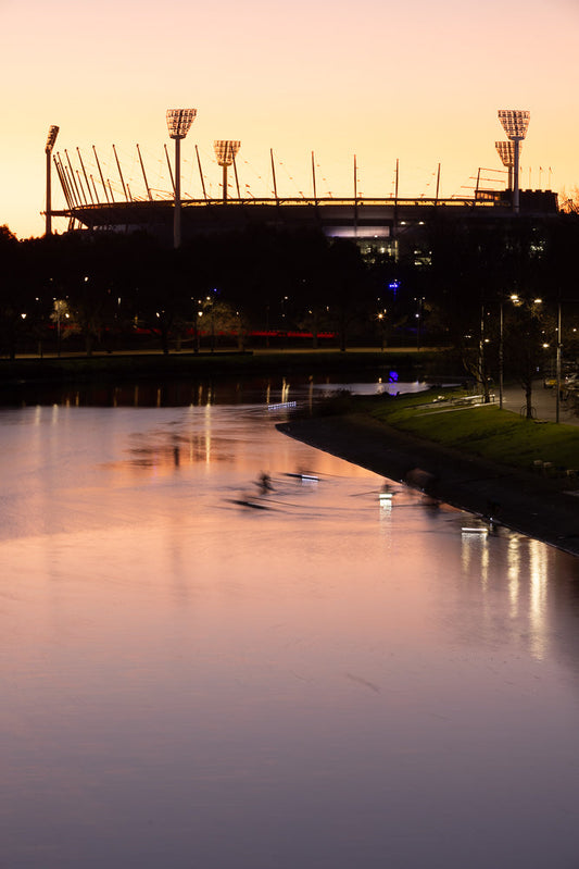 A rowing crew trains on a river at dusk, with the lights of a stadium and city skyline visible in the background under a soft orange and purple sky.