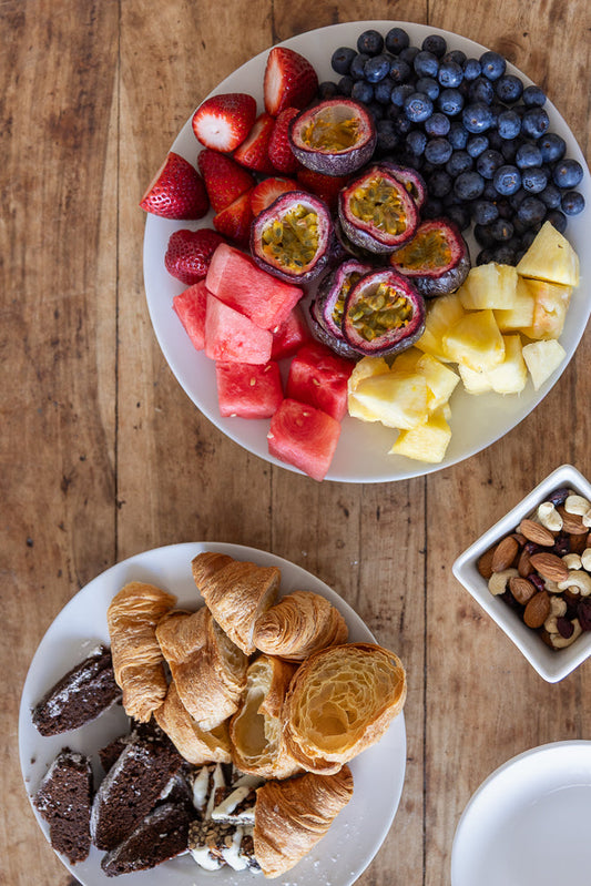 A top-down view of a breakfast spread featuring two white plates on a wooden table. One plate is filled with a colorful assortment of fresh fruits, including strawberries, blueberries, passion fruit halves, pineapple chunks, and watermelon cubes. The second plate holds a variety of pastries, such as croissants and slices of chocolate cake, with a small white bowl of mixed nuts and dried fruit nearby.