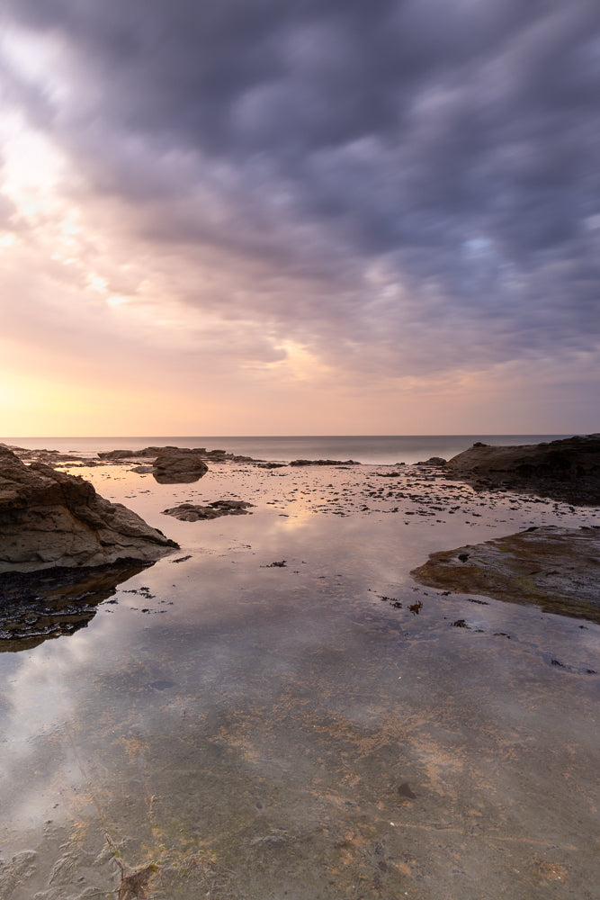 A serene seascape captures the dramatic interplay of light and shadow during a sunset. The sky is a canvas of soft pastels, transitioning from warm orange and pink hues