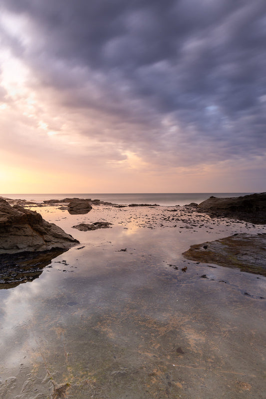 A serene seascape captures the dramatic interplay of light and shadow during a sunset. The sky is a canvas of soft pastels, transitioning from warm orange and pink hues