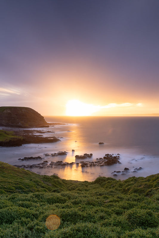 A serene coastal landscape at sunset. The sun, a bright orb, casts a golden glow across the calm ocean, reflecting on the water's surface. Jagged rocks emerge from the misty sea, creating a dramatic contrast with the smooth, blurred water. Lush green grass covers the foreground hills, leading the eye towards the tranquil scene.