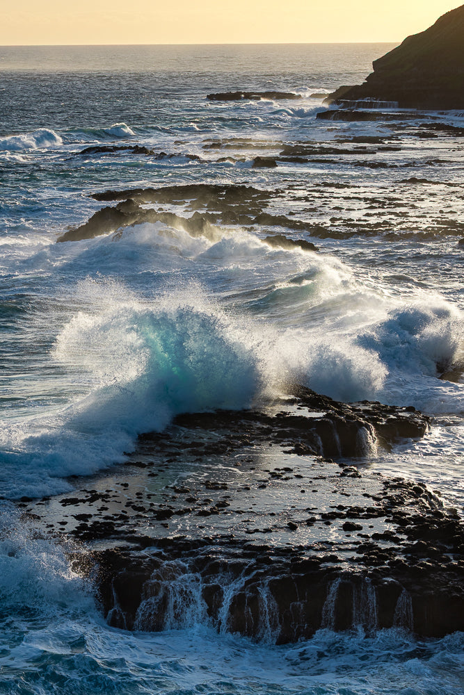Powerful waves crash against dark, rocky shores under a golden sky. The water churns with white foam, creating a dynamic and dramatic seascape.