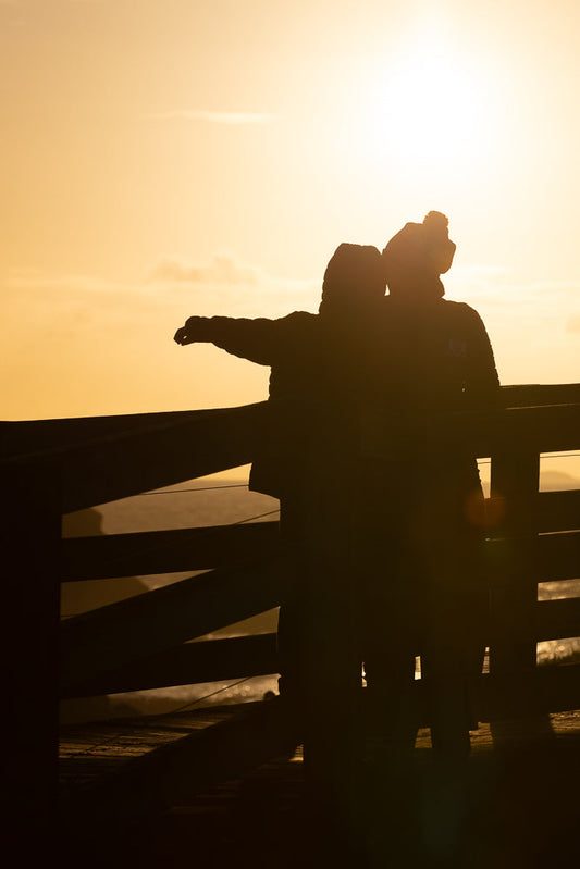 Two people silhouetted against a bright orange sunset, standing on a wooden walkway. One person has their arm outstretched towards the horizon.